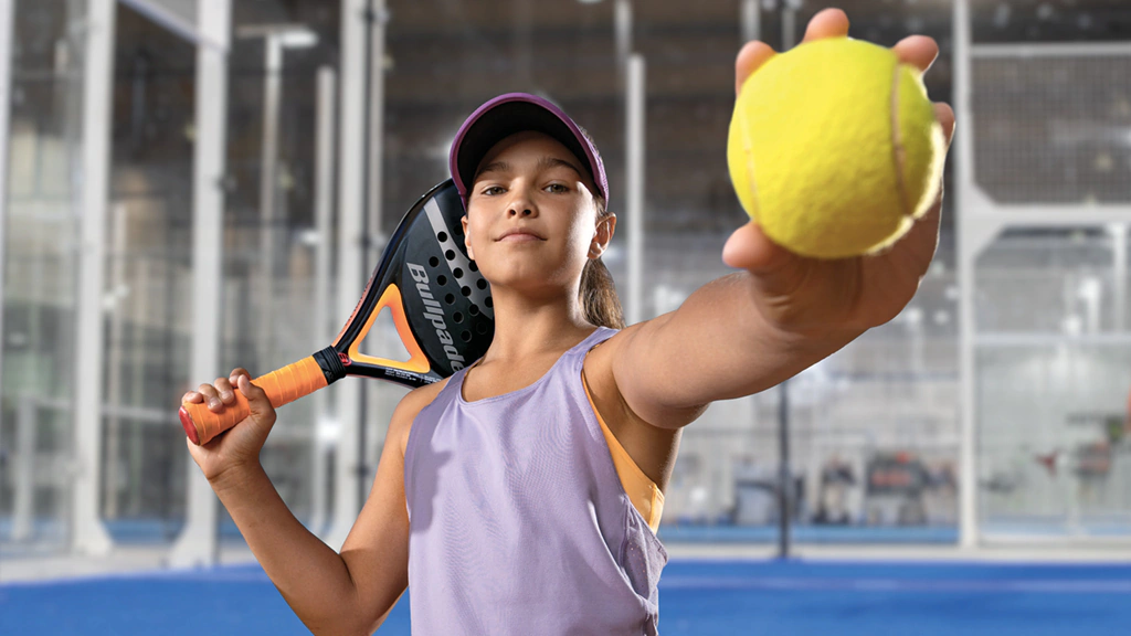 Noia posant amb una pilota i una pala de pàdel en una pista indoor